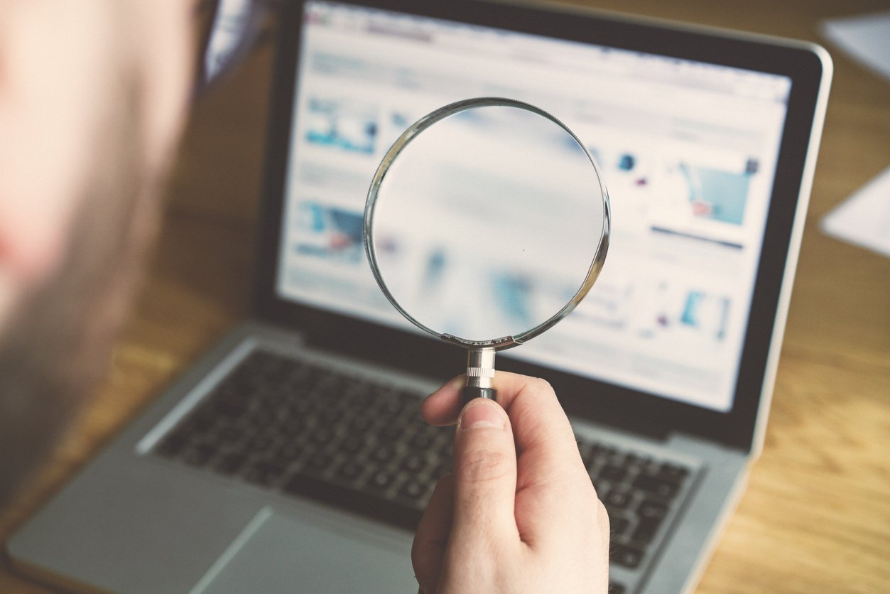 A businessman holding a magnifying glass while looking at his laptop for prospective customers on the internet.